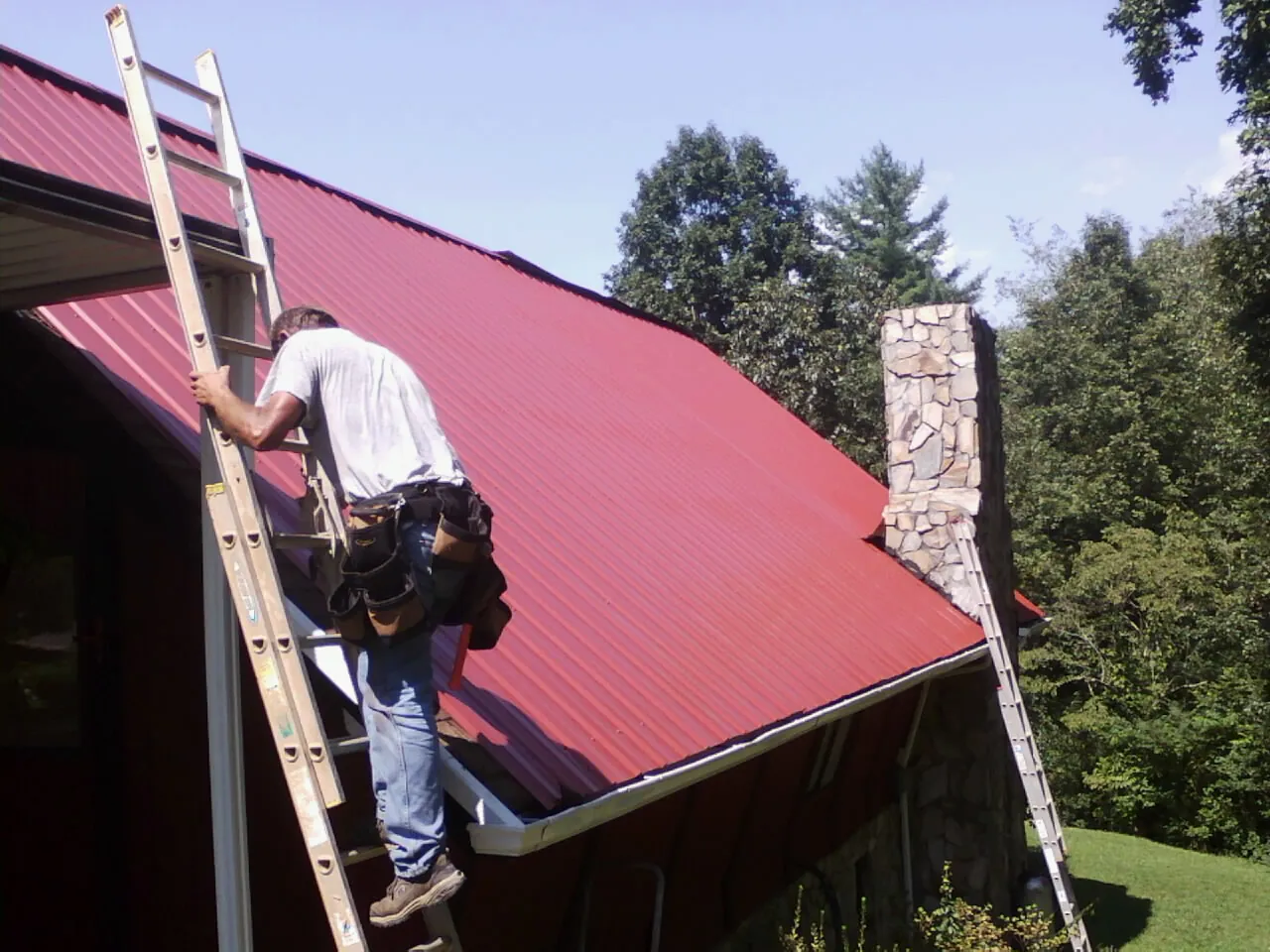 Roofer on a ladder beside a red metal roof and stone chimney — Soffit & Fascia Repair by Roofing Brothers in Everett